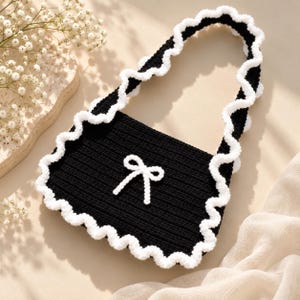May include: A black crocheted handbag featuring a white crocheted bow centered on the front. The bag has a single shoulder strap and is finished with a decorative white crocheted ruffled trim along the entire outer edge of the bag.