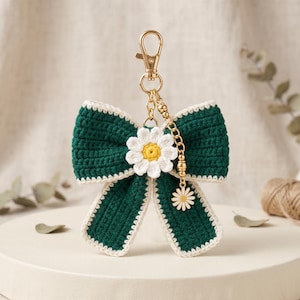 May include: A dark green crocheted bow keychain featuring a white and yellow daisy flower at the center. The bow has a cream-colored border and is attached to a gold-toned metal lobster clasp with a short chain featuring a small daisy charm.