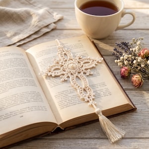 May include: An off-white crochet bookmark featuring an intricate cross-shaped design with delicate lace-like patterns and a long, matching tassel at the bottom, resting flat on the open pages of a vintage book with visible printed text.