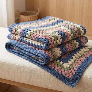 May include: A folded crochet blanket featuring a classic granny square pattern in shades of dusty rose, sage green, cream, and deep blue. The blanket is constructed with textured yarn stitches and finished with a solid blue border along the edges.