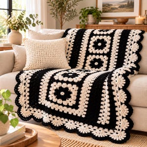 May include: A handmade crochet throw blanket featuring a black and cream granny square pattern with floral motifs and a scalloped edge, draped over a sofa alongside a matching cream-colored square throw pillow with a textured stitch.
