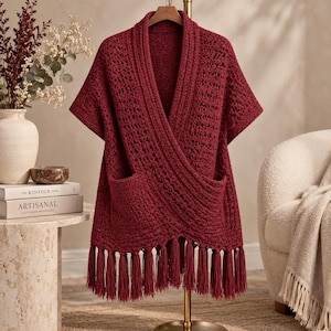 May include: A deep red, short-sleeved, open-front crochet cardigan featuring a textured, lacy stitch pattern, a wide ribbed collar, two front patch pockets, and a bottom hem finished with long, dangling yarn tassels hanging from a wooden hanger.