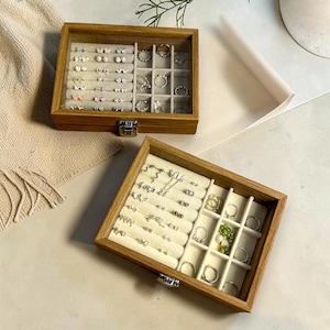 May include: Two rectangular wooden jewelry boxes with clear glass lids and metal latches. The interiors feature cream-colored velvet inserts with multiple rows for earrings and square compartments for rings, currently holding various silver-toned jewelry.