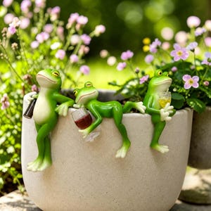 Puede incluir: Tres figuras de ranas verdes posadas en el borde de una maceta de color beige claro. La rana de la izquierda sostiene una botella oscura, la del centro está reclinada con una copa de vino tinto y la de la derecha sostiene un vaso amarillo.