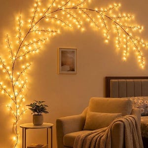 May include: A decorative branch-shaped string light fixture with numerous small, warm-toned LED bulbs arranged in an arching pattern against a wall, featuring thin, dark brown stems that branch out to create a glowing, tree-like silhouette.