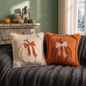 May include: Two square throw pillows featuring a textured, raised pumpkin design with a bow. One pillow is cream-colored with an orange pumpkin, and the other is orange with a cream-colored pumpkin. Both have fuzzy corner accents.