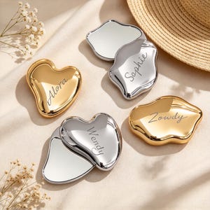 May include: Four compact mirrors with irregular, organic shapes in gold and silver finishes. Each mirror features a custom engraved name: Alora on a gold heart, Sophie on a silver piece, Wendy on a silver piece, and Zowdy on a gold piece.