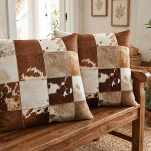 May include: Two square throw pillows featuring a patchwork design of brown and white cowhide squares. The pillows display a variety of natural animal hide textures and patterns, including solid brown, white, and mottled brown and white patches.