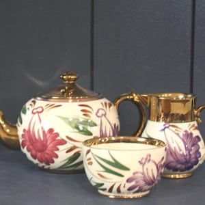 Peut inclure: Un service à thé en céramique composé d'une théière, d'un pot à lait et d'un sucrier. Chaque pièce présente une base couleur crème avec des motifs floraux peints à la main dans des tons rouge, violet et vert, rehaussés d'une finition dorée.
