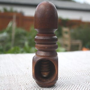 May include: A dark brown wooden nutcracker featuring a rounded top, a series of horizontal decorative rings along the centre, and a central circular opening that reveals a threaded screw mechanism for cracking nuts within the solid wood body.