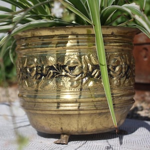 May include: A round brass planter featuring a wide central band with embossed floral and leaf patterns against a dark background. The vessel has horizontal ridged detailing near the rim and base, and it stands on three small, sturdy metal feet.