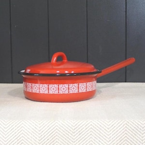 May include: A bright orange enamelled metal saucepan with a matching lid and a long handle. The exterior of the pan features a repeating white geometric pattern around the centre. The rim of the lid and the top edge of the pan are trimmed in black.