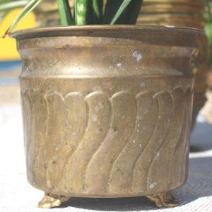 Peut inclure: Un cache-pot cylindrique en laiton orné d'un motif répétitif de vagues verticales en relief sur tout l'extérieur. Le récipient présente un bord large et lisse et repose sur trois petits pieds décoratifs. La surface métallique est patinée.