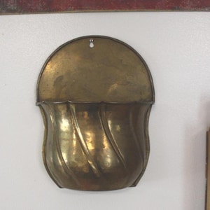 May include: A wall-mounted brass pocket with a semi-circular backplate and a rounded, swirled front section. The metal has a polished, aged finish with visible patina and a single mounting hole at the top centre of the flat back panel.