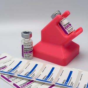 Puede incluir: Dos viales de vidrio con tapas plateadas, uno de pie y otro en un soporte de plástico rosa brillante. Las etiquetas dicen Tirzepatide 20 mg y Retatrutide 10 mg. Una hoja de etiquetas adhesivas con texto de Tirzepatide y GHK-Cu está al frente.