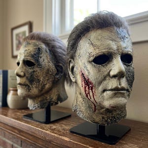 May include: Two realistic latex masks featuring a weathered, pale human face with dark eye sockets, textured skin, and brown hair. One mask displays a prominent red blood-like wound on the right cheek, while both are mounted on black stands.