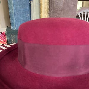 May include: A deep magenta felt hat featuring a wide, matching tonal grosgrain ribbon band encircling the crown. The hat has a structured, rounded top and a flat brim, showcasing a solid, uniform color throughout the felt material and ribbon.
