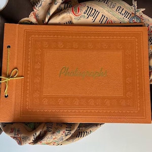 May include: A rectangular orange photo album with a textured cover featuring an embossed border and the word Photographs printed in gold script. The album is bound with a yellow cord through two holes on the left side and rests on a patterned scarf.