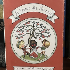 Op de afbeelding: Een hardcover boek getiteld A Year Is Round door Joan Walsh Anglund. De kaft is gedempt rood met een centrale cirkelvormige illustratie van drie kinderen die elkaars hand vasthouden rond een appelboom. De titel en auteur staan op banners.