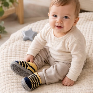 Könnte beinhalten: Ein Paar gehäkelte Babyschuhe mit einem Streifenmuster in Marineblau, Gelb und Grau. Die Schuhe haben eine abgerundete Zehenpartie mit einer einfarbigen grauen Sohle und einem gerippten Bündchen, gefertigt aus strukturiertem Garn für eine flexible Passform.