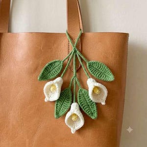 May include: A tan leather tote bag featuring a decorative crochet accessory hanging from the strap. The accessory consists of three white calla lily flowers with yellow centres, surrounded by several textured green crochet leaves on stems.