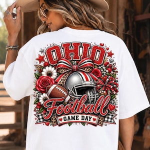 May include: A white t-shirt featuring a graphic on the back with the text OHIO Football GAME DAY. The design includes a football helmet, a football, red roses, white daisies, a cheerleader pom-pom, and a large bow with leopard print accents.