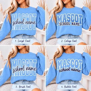 May include: A light blue long-sleeve sweatshirt featuring four variations of the text MASCOT school name in white and dark blue. The designs include block, script, brush, and bubble fonts, showcasing different typography styles for customization.