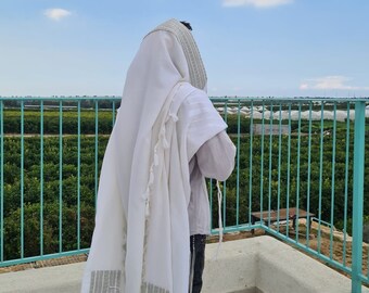 Puede incluir: Una persona con un talit blanco, un chal de oración judío, con rayas grises y borlas. La persona está de pie al aire libre con vistas a un campo verde y un cielo azul.
