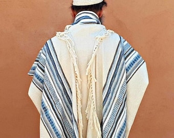 May include: A cream-colored prayer shawl with blue and black stripes and fringed edges. The shawl is draped over a person's shoulders, and a white kippah is worn on the head. The background is a solid, reddish-brown wall.