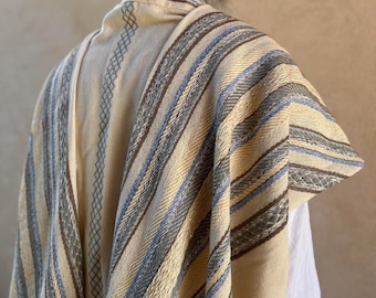 May include: A beige prayer shawl with blue, brown, and gray stripes. The shawl is folded and draped over a person's shoulders.