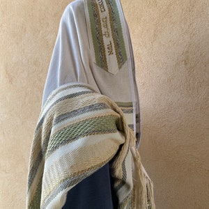 May include: A white prayer shawl with green and gold stripes and a gold embroidered inscription in Hebrew. The shawl is folded and draped over a person's shoulders.