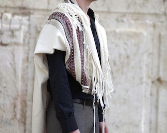 May include: A man wearing a white prayer shawl with burgundy stripes and tassels, standing in front of a stone wall.