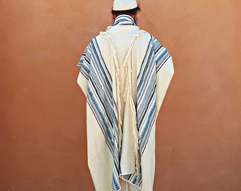 May include: A white prayer shawl with blue and black stripes and fringe. The shawl is worn by a person standing in front of a brown wall.