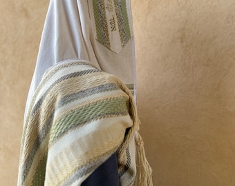 Puede incluir: Una chal de oración blanca con rayas verdes y doradas y borlas. La chal tiene escritura hebrea.