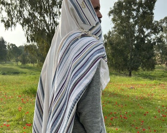 May include: A person wearing a white prayer shawl with blue and gold stripes and the Hebrew text "כבוד" embroidered on the shawl. The person is standing in a field of green grass with red flowers.