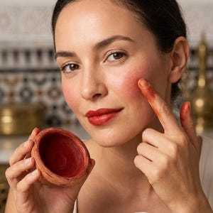 May include: A small, round, terracotta-coloured clay pot containing a deep red pigment. The pot has a textured, earthy exterior and a smooth, circular opening. The red pigment is applied to the cheek and matches the deep red lipstick on the lips.