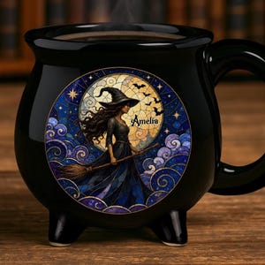 May include: A black ceramic cauldron-shaped mug featuring a circular stained-glass style illustration of a witch with a broomstick against a full moon and starry night sky. The name Amelia is printed in black script on the moon background.