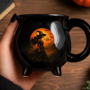 May include: A black ceramic cauldron-shaped mug featuring a graphic of a witch in a hat holding a broomstick against a large orange full moon with flying bats. The name Olivia is printed in black script text on the orange moon background.