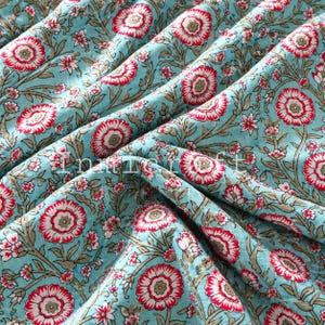 May include: A light blue fabric featuring a repeating floral pattern with red and white petals, green centers, and brown stems. The textile displays a dense botanical print with intricate vines and leaves. The word Innieraft is visible.