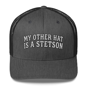 May include: A charcoal grey trucker hat with a black mesh back and a structured front panel. The front features white embroidered text that reads, "MY OTHER HAT IS A STETSON," arranged in two arched lines across the center of the cap.