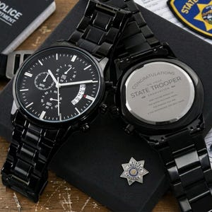 May include: A black stainless steel chronograph watch with a black dial and a metal link band. The back of the watch case is engraved with the text: CONGRATULATIONS ON YOUR STATE TROOPER GRADUATION. MAY YOU BE PROUD OF THE WORK YOU HAVE DONE.