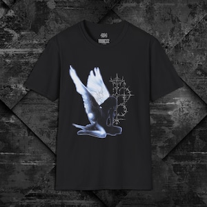 以下が含まれることがあります： 黒の半袖Tシャツで、光る白い翼のある人物が細い棘のある十字架のデザインの横に跪いているグラフィックが描かれています。首元の内側のラベルには、黒い生地を背景に白い文字で404 VAULTZと記されています。