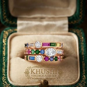Op de afbeelding: Een stapel van drie gouden ringen met diverse veelkleurige edelstenen, waaronder ronde witte diamanten en rechthoekige roze, groene, blauwe en gele stenen. De ringen liggen in een groen fluwelen doosje met de tekst KHUSHI VINTAGE ATELIER.
