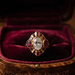 Peut inclure: Une bague en or ornée d'une pierre centrale transparente taille ovale, entourée d'un halo de petits rubis rouges de formes variées, incluant des tailles ovale, carrée et baguette, présentée dans un écrin en bois doublé de velours rouge.