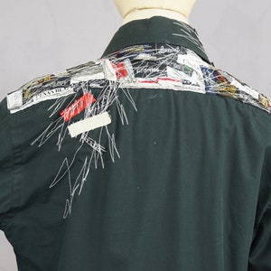 May include: A dark green long-sleeved shirt with a patchwork design on the back. The design features various colorful fabric scraps and white stitching.