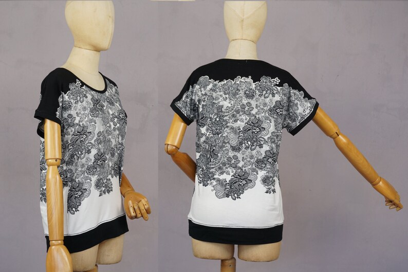 May include: A white and black short-sleeved shirt with a floral print. The shirt has a black band around the neck and a black band at the bottom.