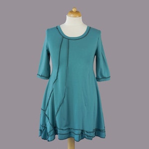 Op de afbeelding: Turquoise blauwe tuniekjurk met een uniek asymmetrisch ontwerp en zwarte stiksels. De jurk heeft een ronde halslijn en 3/4 mouwen.