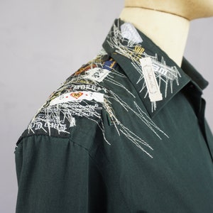 May include: A dark green long-sleeved shirt with white embroidery on the collar and shoulder. The embroidery includes various small fabric scraps and labels with text.