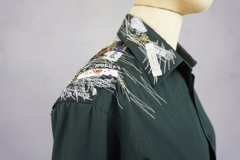 May include: A dark green long-sleeved shirt with white embroidery on the collar and shoulder. The embroidery includes various small fabric scraps and labels with text.
