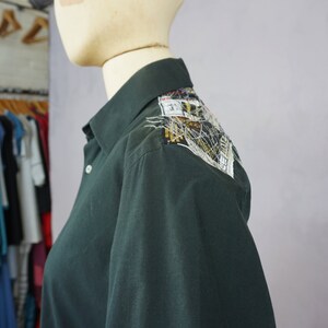 May include: A dark green long-sleeved shirt with a decorative patch on the shoulder. The patch is made of various fabrics and includes a small label with the text "31".